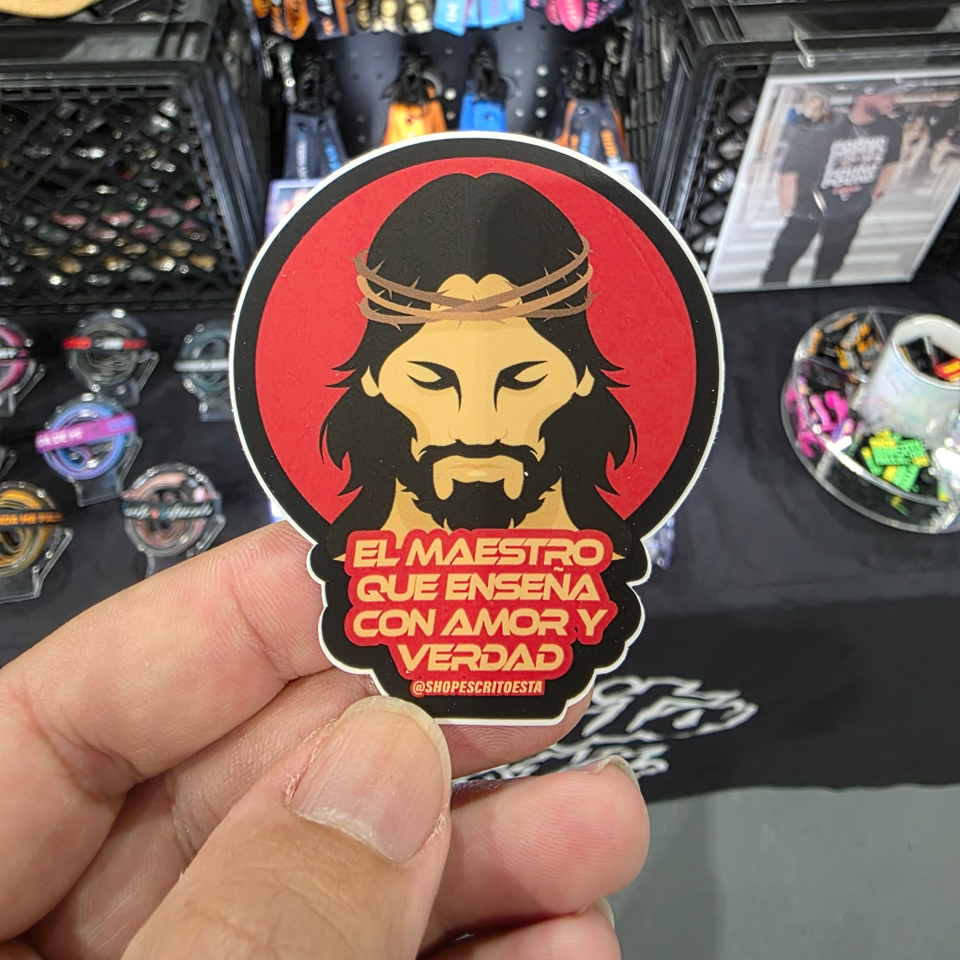 Sticker of a religious figure with text held by a hand, with a blurred background of merchandise.